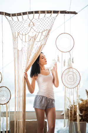 A young woman on a window curtains. Macrame hobby weaving dream catchers, feathers. Girl weaves threadsの写真素材