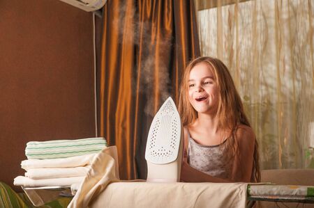 little funny housewife with iron. Iron in the hand of the girl on the Ironing Board irons and steams clothes. Little girl playing with iron. Danger at home. Beauty girl with ironing dressの写真素材