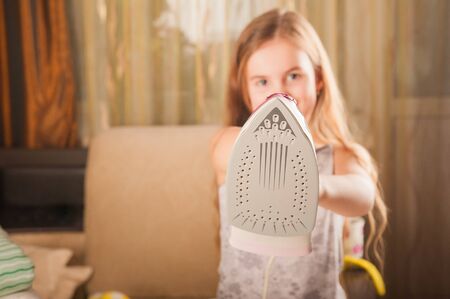 little funny housewife with iron. Iron in the hand of the girl on the Ironing Board irons and steams clothes. Little girl playing with iron. Danger at home. Beauty girl with ironing dressの写真素材