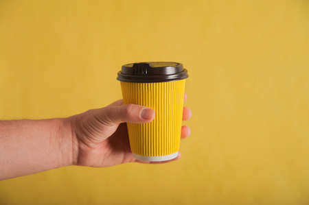 Hand with yellow nails holds a paper cup on a yellow background close-up and copy space. Monochrome...