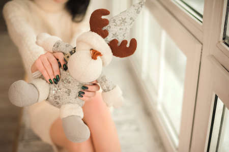 Girl in a white sweater dress against the background of a large window in a rustic style. Young woman at home on the windowsill for Christmas and New Year.の写真素材