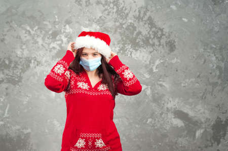 Girl in a Santa Claus hat on a background of a concrete gray wall close-up and copy space. Young woman in a New Year's costume and a face mask from COVID 2019.の写真素材