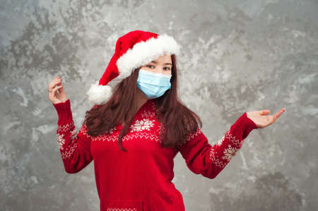 Girl in a Santa Claus hat on a background of a concrete gray wall close-up and copy space. Young woman in a New Year's costume and a face mask from COVID 2019.の写真素材