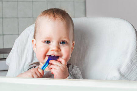 Cheerful child eats food with spoon. Close up portrait of happy boy in high chair. Complementary...