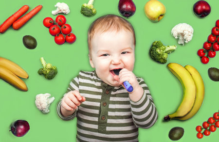 toddler with spoon on green background surrounded by fruits and vegetables. Complementary feeding of...