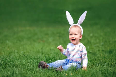 Funny toddler with bunny ears is looking for eggs in grass. Easter egg hunt. Ostern.