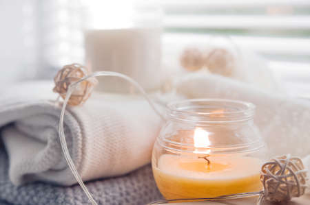 Burning candles and white sweaters on the windowsill. Cozy autumn concept and a window with blinds.の写真素材
