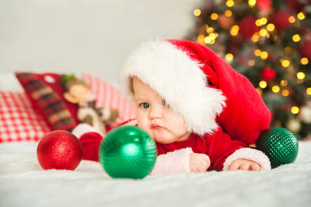 Baby boy dressed as Santa Claus for Christmas plays with wooden train and Christmas decorations in...