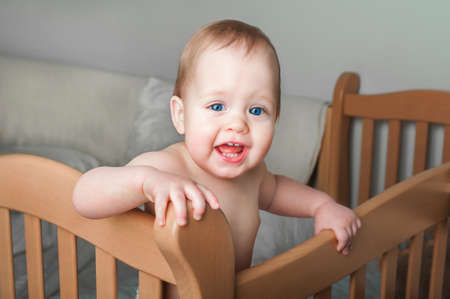 Portrait of beautiful baby 11 months old in diaper on bed close-up. Skincare, healthcare concept for children. Teething, hands on toddler's mouth.の写真素材
