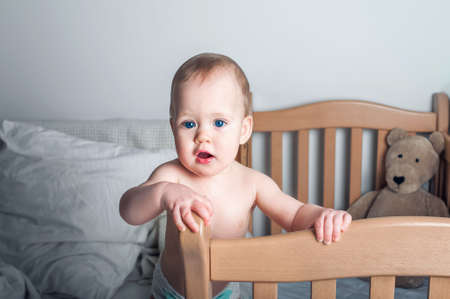 Portrait of beautiful baby 11 months old in diaper on bed close-up. Skincare, healthcare concept for children. Teething, hands on toddler's mouth.の写真素材