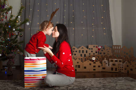 Young happy mom and daughter in red sweaters open gifts in bags at home. Christmas mood, DIY home decor from cardboard.の写真素材