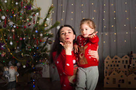 Young happy mom and daughter in red sweaters playing, hugging, fooling around at home. Christmas mood, DIY home decor from cardboard.の写真素材