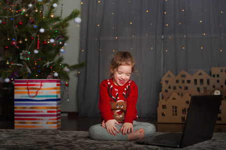 Little girl child in red Christmas sweater for New Year at home with laptop congratulations.の写真素材
