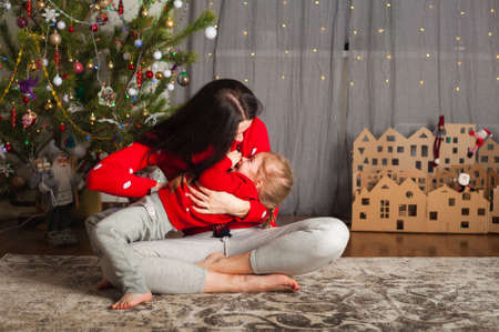 Young happy mom and daughter in red sweaters playing, hugging, fooling around at home. Christmas mood, DIY home decor from cardboard...の写真素材