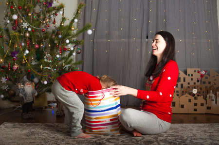 Young happy mom and daughter in red sweaters open gifts in bags at home. Christmas mood, DIY home decor from cardboard...の写真素材