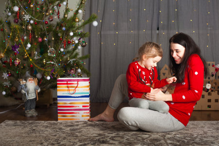 Young happy mom and daughter in red sweaters playing, hugging, fooling around at home. Christmas mood, DIY home decor from cardboard...の写真素材