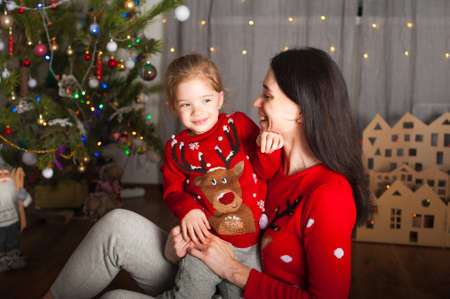 Young happy mom and daughter in red sweaters playing, hugging, fooling around at home. Christmas mood, DIY home decor from cardboard.