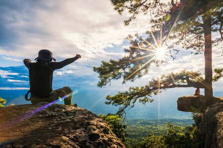 man Sitting on stone at the Cliff edge on top of mountain watching the sunset, travel and freedom conceptの写真素材