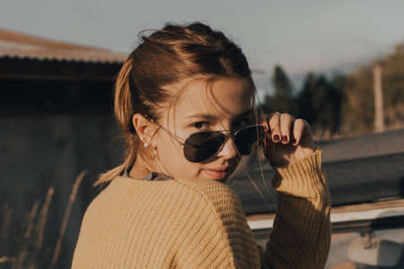 beautiful young girl looking at the camera sitting on an old car at sunset.の写真素材