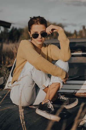 beautiful young girl looking at the camera sitting on an old car at sunset.の写真素材