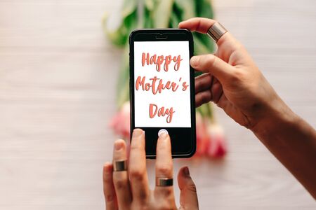 happy mother's day text sign on screen of phone. hand holding gadget with message   above pink tulips on white wooden rustic background. greeting card conceptの写真素材