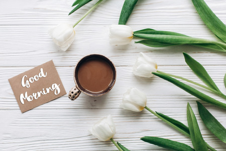 good morning text sign on greeting card at tulips with coffee on white wooden rustic background with space for text. stylish flat lay with flowers and drink. hello springの写真素材