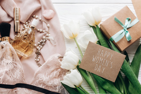 happy womens day text sign on woman lace lingerie jewelry and perfume present on soft fabric and tulips with empty greeting card on white rustic background. flat layの写真素材