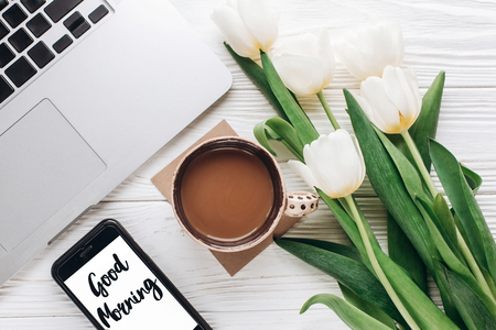 good morning text sign on phone screen and laptop with morning coffee and tulips on white wooden rustic background. stylish flat lay with flowers and working gadgets. freelance. home workingの写真素材