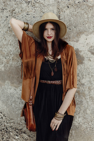 stylish boho woman with jewelry posing at rock wall. beautiful hipster gypsy dressed girl with hat and fringe poncho with sensual look. young girl traveler. fashionable hippie outfit. summer vacationの写真素材