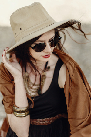 beautiful stylish hipster woman in sunglasses holding hat, with windy hair. boho traveler girl in gypsy look, near beach in mountains. atmospheric moment. summer travel. space for text.の写真素材