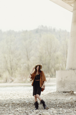 beautiful stylish boho woman smiling, with hat, fringe poncho. girl in gypsy hippie look young traveler posing near river rocks in mountains. sensual look. atmospheric moment. summer travelの写真素材