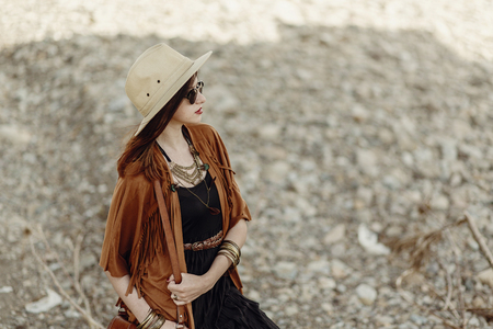 beautiful stylish boho woman in sunglasses and hat, fringe poncho and leather hat. hipster girl in gypsy look young traveler posing near river beach in mountains.  atmospheric momentの写真素材