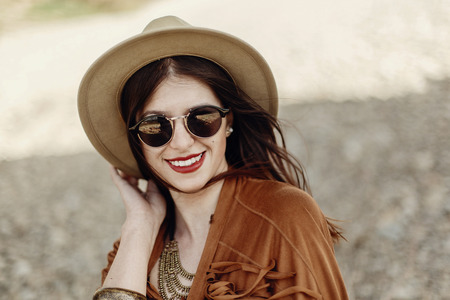 stylish boho woman smiling in sunglasses holding hat, with windy hair. hipster girl in gypsy look, young traveler near river beach in mountains. atmospheric moment.  space for text.の写真素材