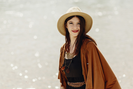 stylish boho traveler woman in hat, fringe poncho posing near water river beach, gypsy hipster girl look, summer travel. atmospheric moment. space for text. wanderlustの写真素材