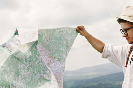 stylish hipster traveler man holding map and exploring at top of mountains with amazing view, summer...