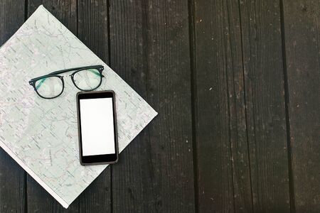 stylish phone with empty screen and glasses on map on black wooden background, space for text, travel wanderlust concept, application mock-upの写真素材