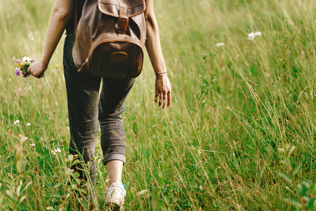stylish hipster woman walking in grass and holding  in hand herb  wildflowers  in summer mountains, travel concept, peaceful relaxing momentの写真素材
