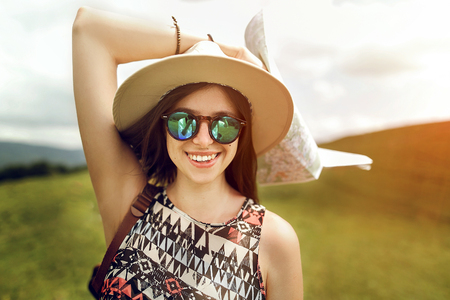 stylish hipster woman traveler holding map, with fashionable sunglasses and hat smiling on top of mountains in summer, travel concept, space for textの写真素材