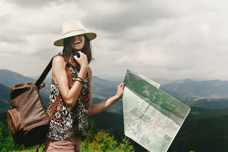 stylish hipster woman traveler with backpack holding map and laughing with hat on eyes on top of mountains, travel  concept, space for textの写真素材