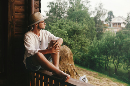 hipster man sitting on porch of wooden house  with field and hay in evening, calm moment, summer farm in countryside concept, space for textの写真素材