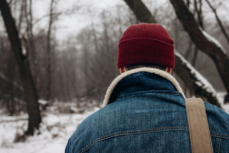 stylish hipster traveler with backpack in red hat walking in winter snowy forest. wanderlust and adventure concept with space for text. back view. atmospheric momentの写真素材