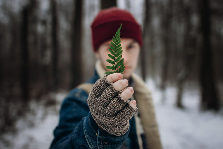 stylish hipster traveler holding green leaf fern in front of face, save the earth concept. environment protection. wanderlust and adventure concept in winter forest.の写真素材