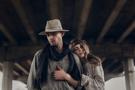 stylish hipster couple dreaming under abandoned bridge. boho gypsy woman gently hugging arm of confident man in hat. atmospheric sensual moment. rustic fashionable look.の写真素材