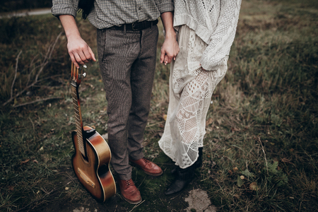 stylish hipster couple holding hands closeup in windy field. boho gypsy woman and man with guitar  hugging. atmospheric moment. fashionable look. rustic wedding conceptの写真素材