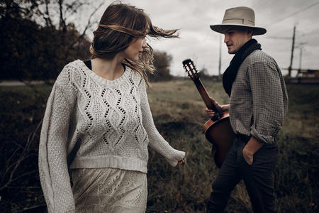 stylish hipster couple posing. man in hat with guitar and his boho woman in knitted sweater in windy field. atmospheric sensual moment. fashionable look. rustic wedding conceptの写真素材