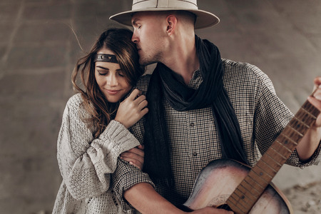 stylish hipster couple gently hugging. man in hat playing guitar for his boho woman in knitted sweater. atmospheric sensual moment. rustic fashionable look.の写真素材
