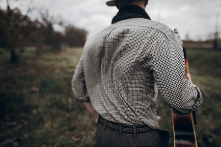 stylish hipster man back in windy field with guitar walking to his woman. atmospheric moment. fashionable look. rustic wedding conceptの写真素材