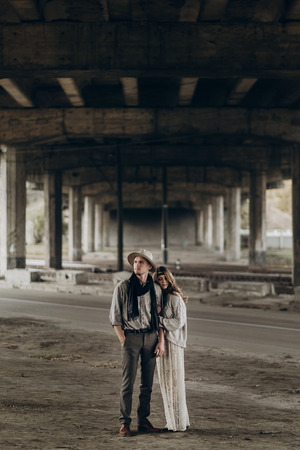 stylish hipster couple gently hugging. man in hat and boho woman in knitted sweater under abandoned bridge. atmospheric sensual moment. rustic fashionable look.の写真素材