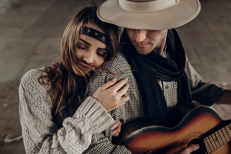 Romantic brunette hipster girl in boho clothes hugging handsome cowboy musician with guitar and a scarf closeupの写真素材