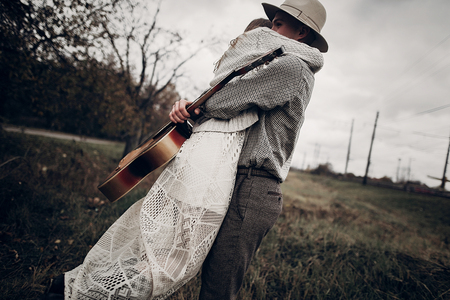 Stylish hipster couple hug in field, handsome cowboy musician with guitar and indie style woman in white sweater pose near railway stationの写真素材